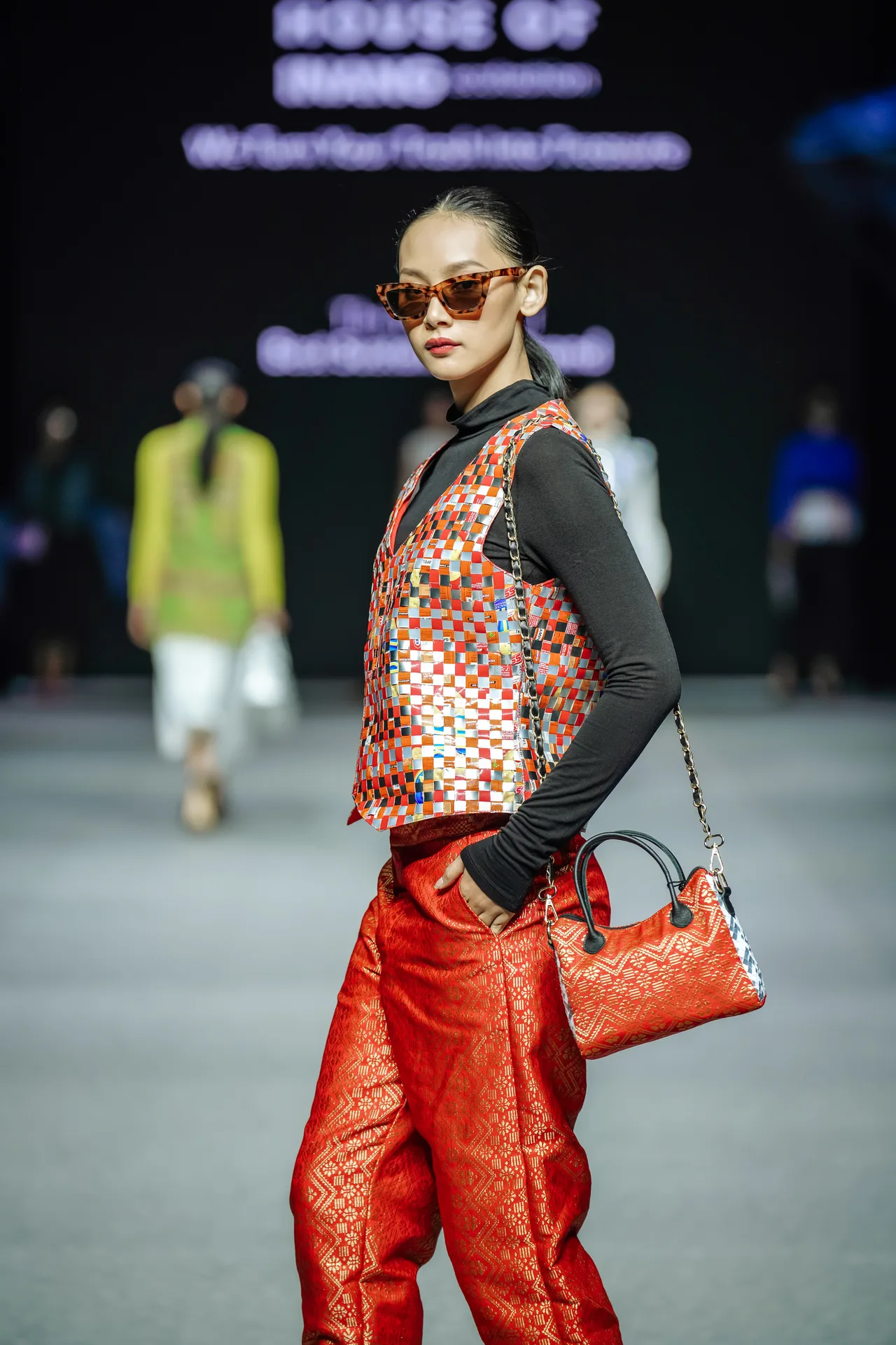 Runway model wearing orange sunglasses, woven checkered vest, red satin trousers and handbag