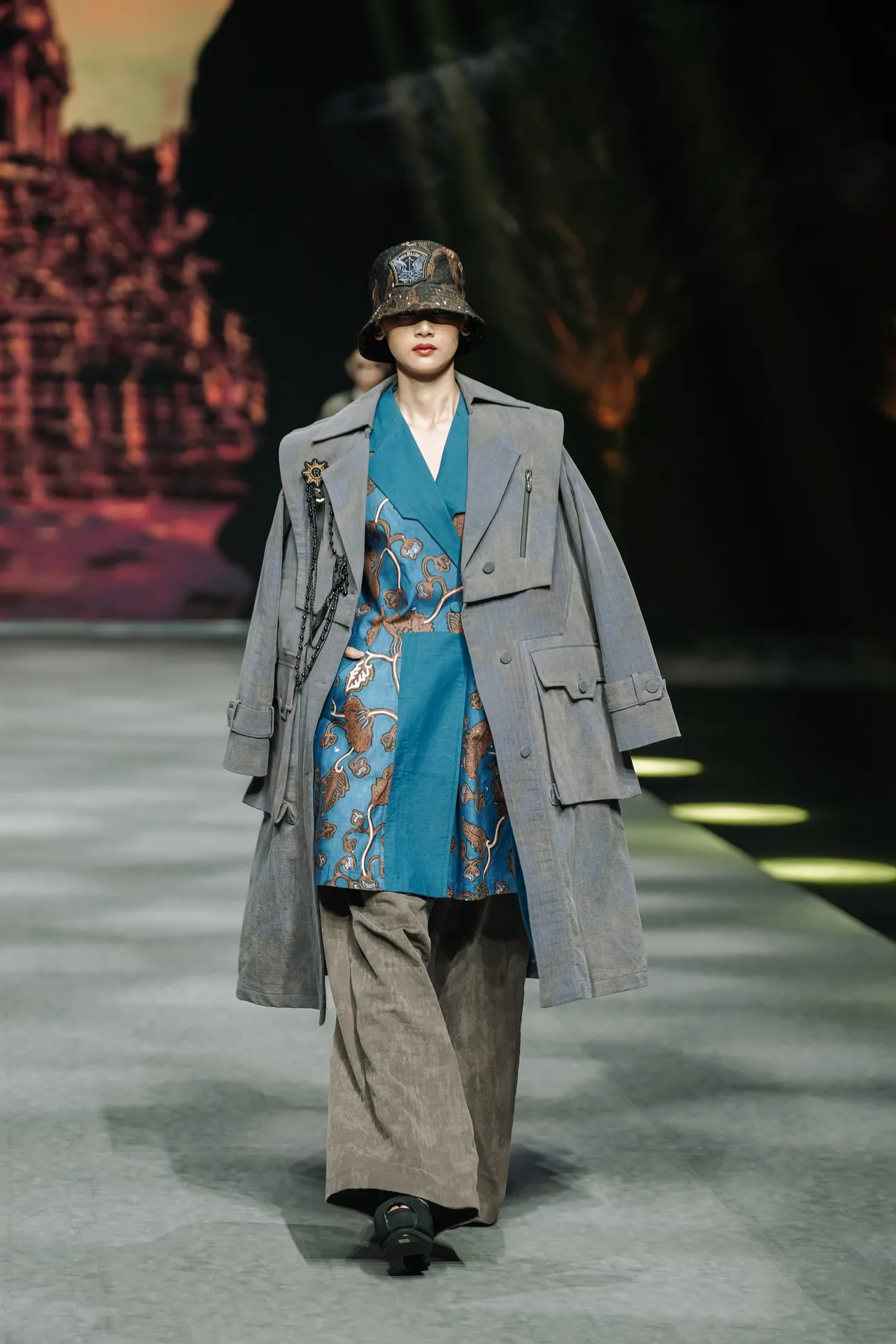 Full runway walk of model in patterned bucket hat, grey trench coat over turquoise vest and wide-leg trousers