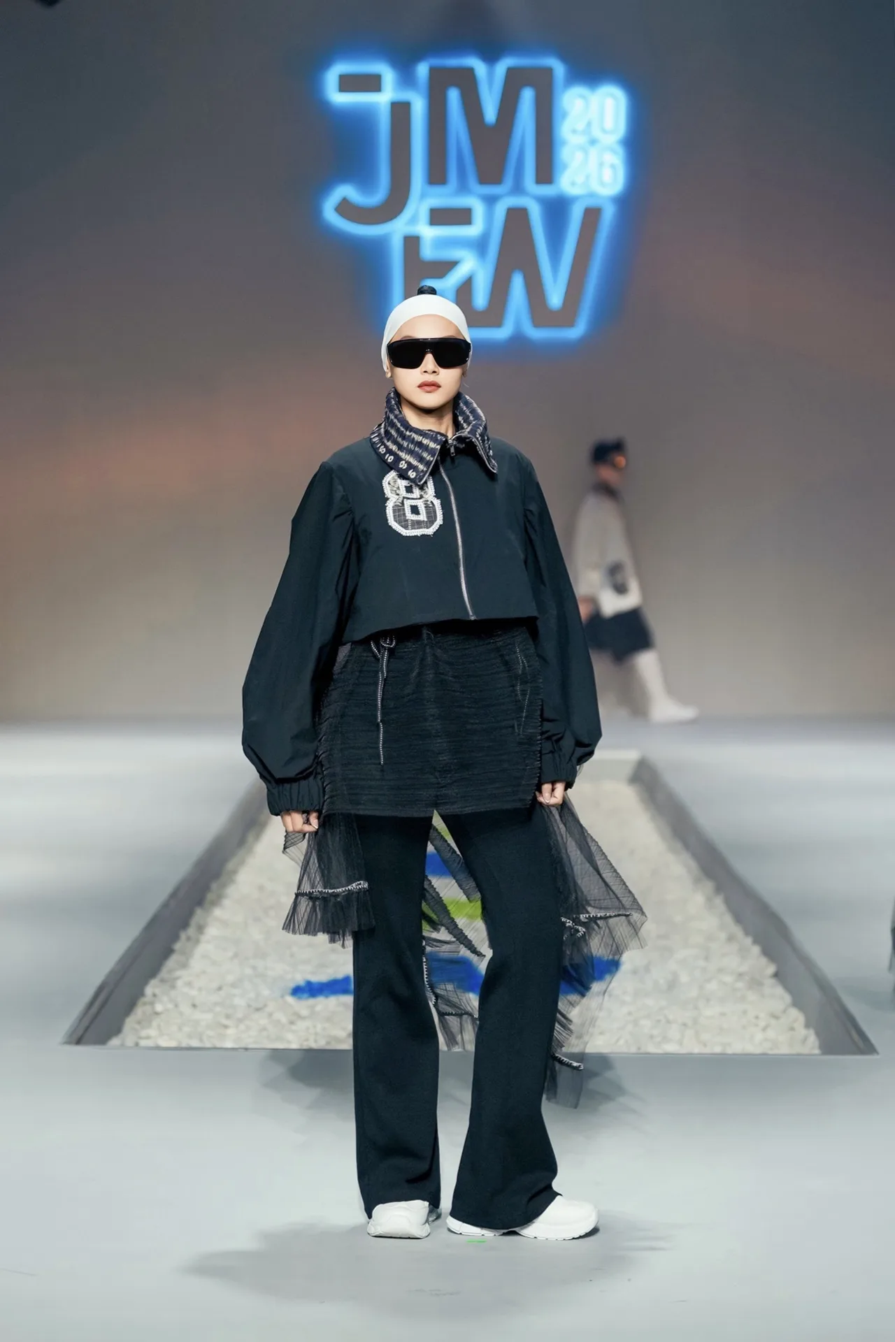 Full body runway walk of model in black sporty jacket with sheer tulle trim and flared trousers at JMFW